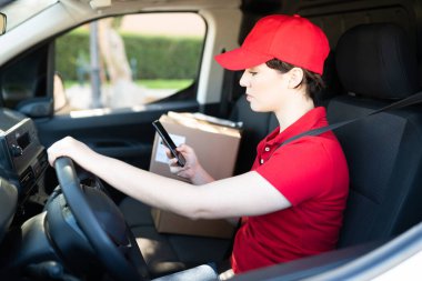 20 'li yaşlarda beyaz bir kadın teslimat kamyonunda şoför olarak çalışıyor. Dişi kurye akıllı telefonuna bakıyor ve bir sonraki paketi teslim etmek için bilgi okuyor.