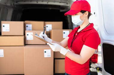 Paketi teslim etmek için bir panoya bilgi ve nakliye adresi yazan maske ve eldivenli çekici bir teslimatçının yan görüntüsü.