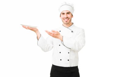 Beyaz üniformalı neşeli şef tabakta yeni lezzetli bir yemek gösteriyor. Güzel genç adam bir restoranda aşçı olarak çalışıyor ve akşam yemeği için lezzetli bir yemek hazırlıyor.