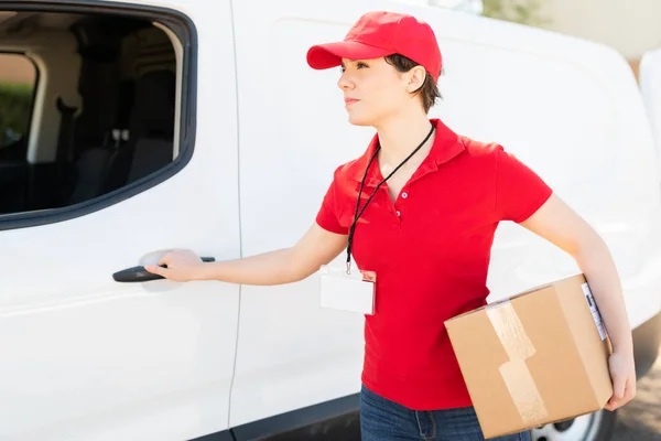 Caucasian delivery woman carrying a small box after picking up some packages to deliver at a shipping address. Female courier opening the door of a delivery van