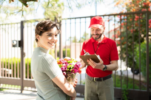 Portrait of a beatiful young woman smiling after receiving a bouquet of flowers in her doorstep. Delivery man holding a clipboard outside a woman\'s house