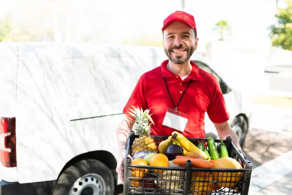 Happy delivery man in his 30s smiling while holding a box full of beautiful vegetables and fruits. Latin male worker carrying a package for a delivery service