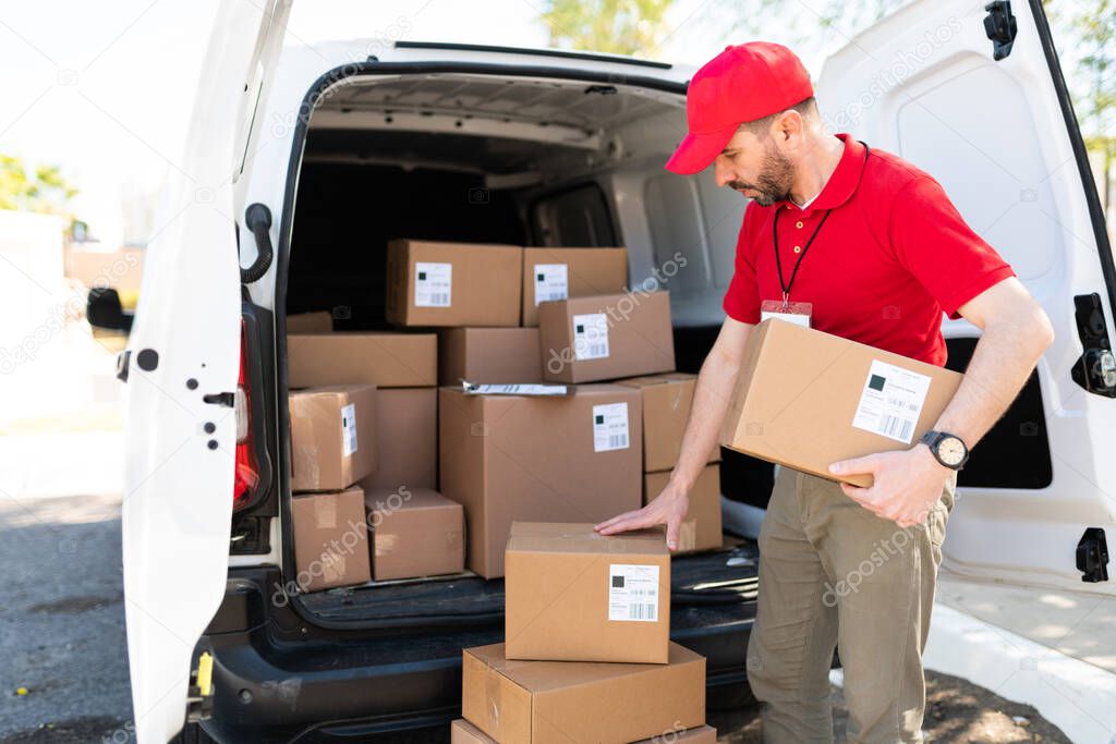Male worker in a red uniform unloading the packages and parcels of a delivery van. Hispanic courier delivering boxes at a home or business shipping address 