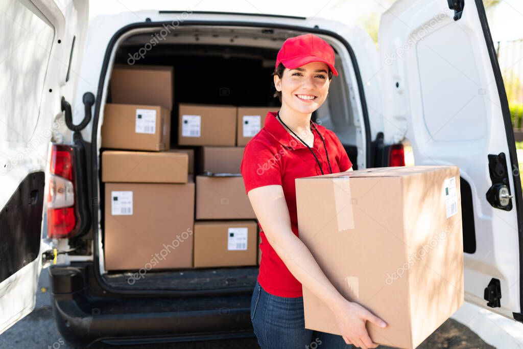 Portrait of an attractive caucasian female worker in a red uniform unloading packages while smiling and standing next to a delivery van with a lot of boxes and parcels