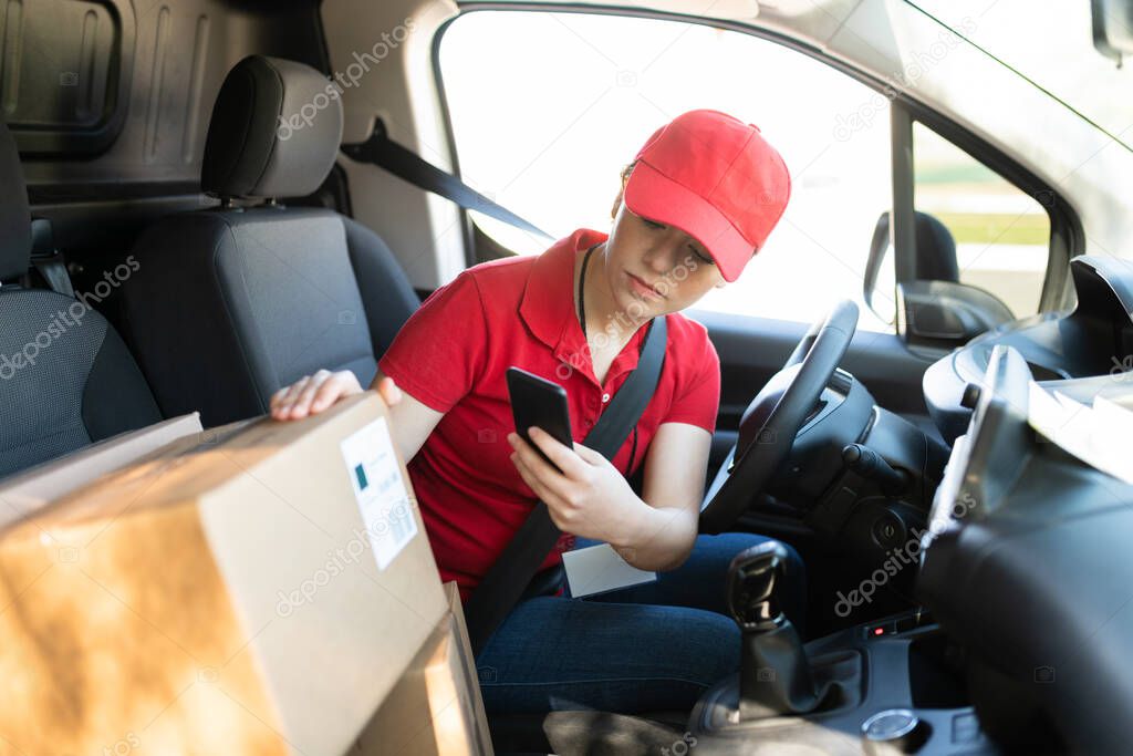 Caucasian young woman and driver scanning the barcode of a shipping label of a package while sitting behind the wheel of a delivery van before delivering the parcel
