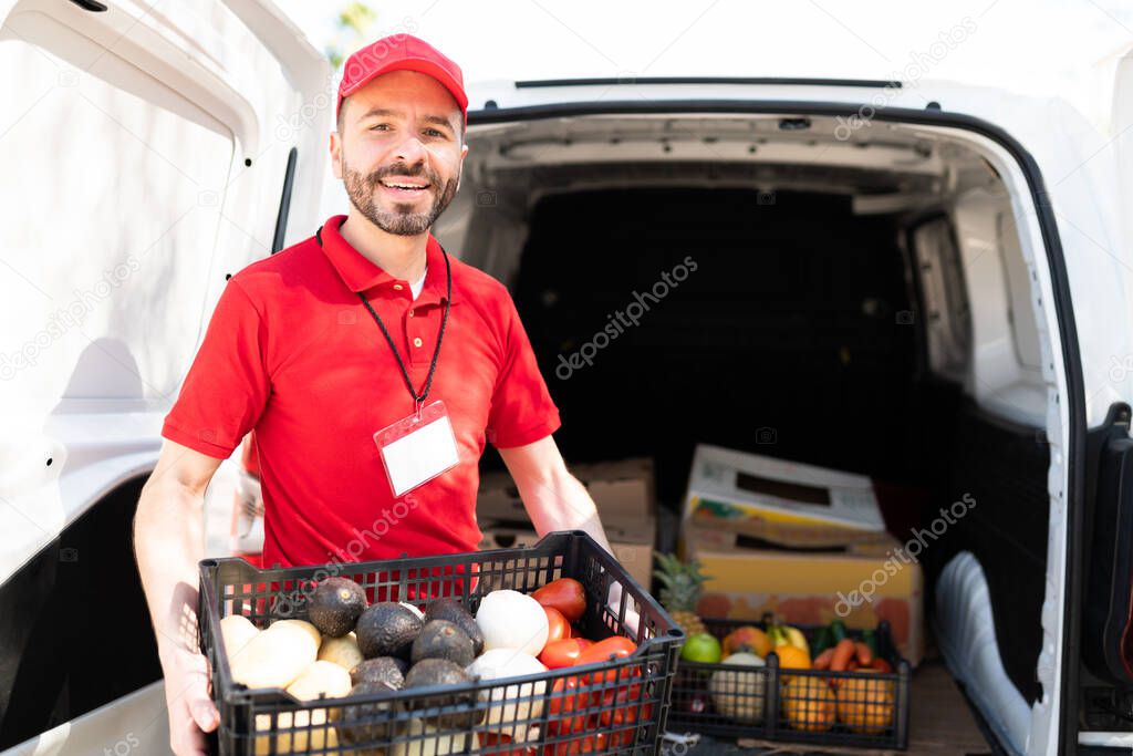 Cheerful delivery man holding a box of fresh fruits and vegetables in the back of a white truck. Male worker from a supermarket making a delivery at home