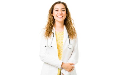 Göz teması kurarken beyaz önlük ve steteskop takan mutlu İspanyol doktor. Beyaz bir arkaplan önünde gülümseyen ve poz veren profesyonel bir genç kadın.
