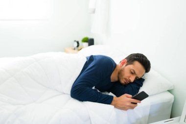 30 'lu yaşlarda çekici bir adam yatakta uzanıyor ve akıllı telefondan sosyal medyayı kontrol ediyor. İspanyol adam yatak odasında dinlenirken mesaj atıyor.