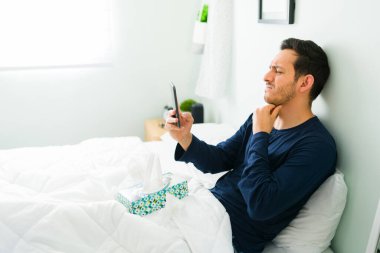 Hispanik bir adam kendini kötü hissediyor ve akıllı telefonuyla çevrimiçi video görüşmesi sırasında sanal bir doktorla konuşuyor. Ateşi olan ve boğazı ağrıyan bir adam yatakta yatıyor.