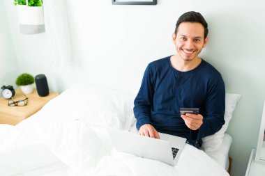 Mutlu adam pijamalarıyla yatakta otururken gülümsüyor ve göz teması kuruyor. Elinde kredi kartı olan yakışıklı bir adam ve online alışverişini laptoptan ödemeye hazır. 