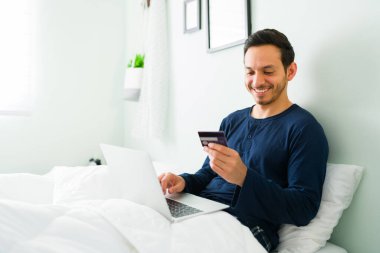Pijamalı gülümseyen Latin adam online alışverişini kredi kartıyla ödüyor. İnternetten bilgisayar alan yakışıklı bir adam. 