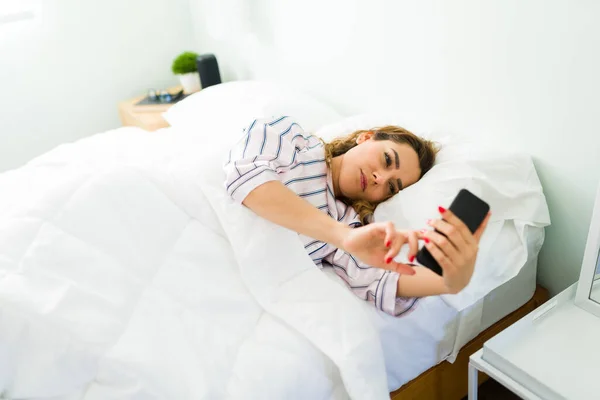 ラテン女性で彼女の30代のベッドで寝そべって後に目が覚めた彼女のテキストと仕事の電子メールオンザスマートフォン — ストック写真
