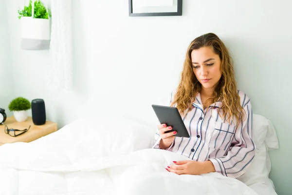 美しい女性はピンクのパジャマを着て タブレットや電子リーダーと朝のコーヒーカップで本を読んでいる間 彼女の快適な白いベッドの上に座っています — ストック写真
