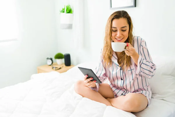 快乐的女人穿着睡衣躺在床上 边喝咖啡边阅读平板电脑上的电子书 — 图库照片