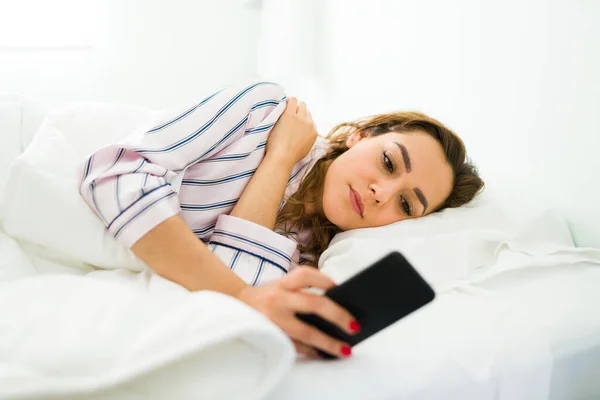 三十多岁的迷人女人躺在床上 用智能手机查看社交媒体 西班牙裔女人在卧室休息时发短信 — 图库照片