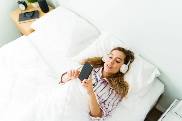 30代のリラックスした女性の高い角度で ヘッドフォンがベッドに横たわっていると彼女のスマートフォンでテレビシリーズを見て — ストック写真