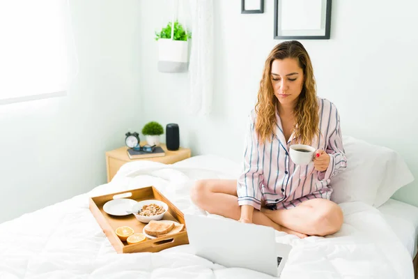 迷人的女人在笔记本电脑上打字 坐在床上工作 拉丁美洲女人在床上吃早餐 — 图库照片