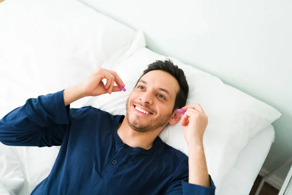 Smiling young man in pajamas putting on ear plugs before sleeping on her comfy bed to avoid the noise