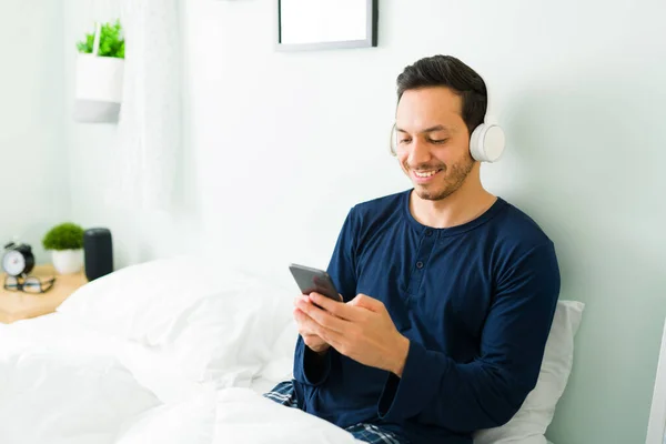 兴奋的拉丁男人一边在白色的床上休息 一边用耳机听音乐 快乐的男人喜欢用他的智能手机唱歌 — 图库照片