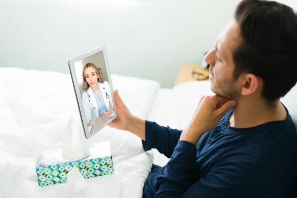 一位英俊的男人在床上休息时 在平板电脑上与一位虚拟医生在线视频通话 抱怨疼痛和嗓子疼 — 图库照片