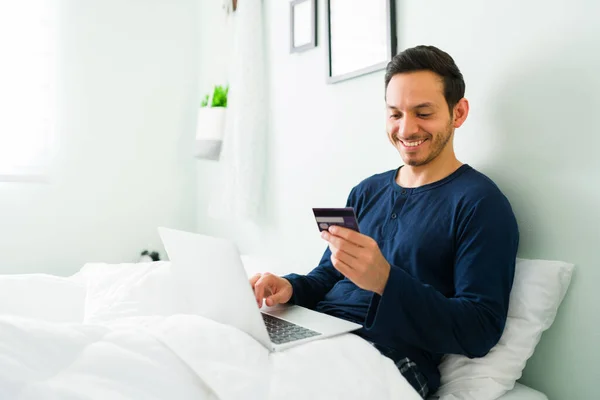 Smiling latin man with pajamas paying for his online shopping with a credit card. Good-looking man using his laptop to buy online