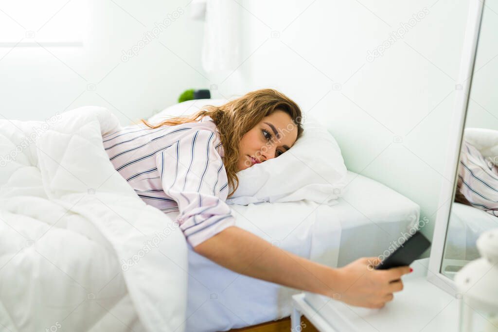 Tired attractive woman checking the late time in her smartphone while lying and resting on her stomach in bed