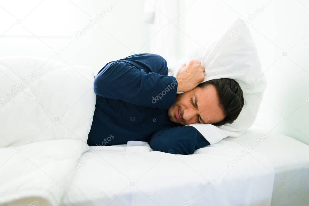 Upset latin man lying in bed and covering his ears with a white pillow because of the loud noise 