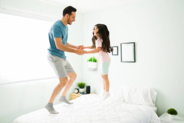 Çekici genç baba ve pijamalı küçük kız beyaz rahat bir yatakta zıplıyorlar. Latin bir adam ve kızı yatak odasında oynarken gülüyorlar.