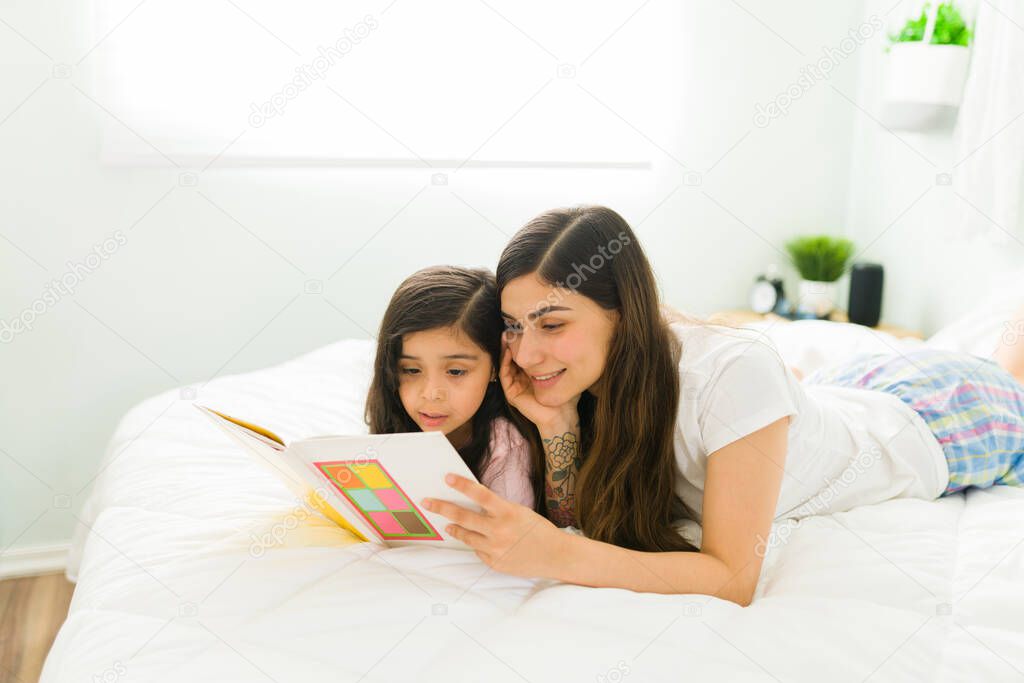 Happy mother smiling while reading to her little daughter a fairy tale. Beautiful mom and cute girl relaxing with a children's book in bed