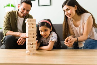 Tahta oyun kulesinin tahta bloğunu çekip hamlesini yapmaya çalışan odaklanmış küçük kız. Latin ebeveynler kızıyla eğleniyor.