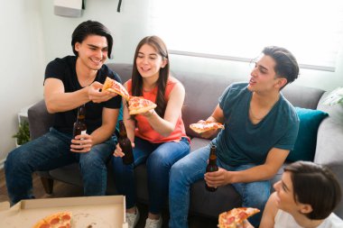 Bir grup genç erkek ve kadın kanepede oturmuş üniversiteli arkadaşlarıyla küçük bir toplantı sırasında lezzetli pizzalar yiyorlar.