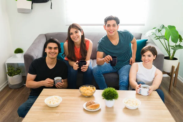 Portrait of young college friends smiling and making eye contact while hanging out in the living room with coffee and snacks