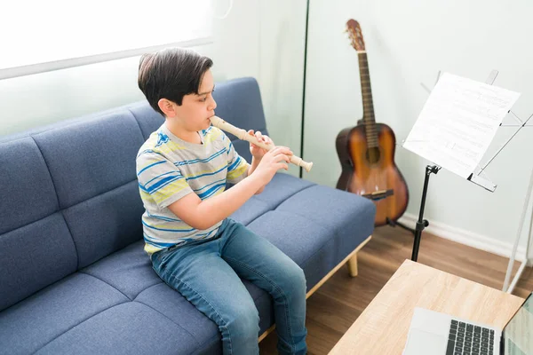 音楽のスキルを磨く 白人の小学生は 彼のオンラインレッスンのためにリビングルームに座っている間 フルートで自分の好きな曲を演奏 — ストック写真