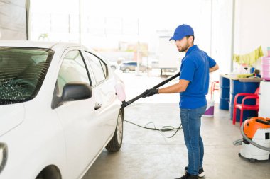 Otomobil bakım servisinde çalışan İspanyol bir çalışan. Üniformalı genç adam araba yıkamada çalışıyor. Erkek işçi su hortumuyla bir aracı yıkıyor. 