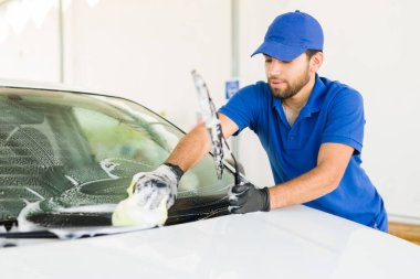 Sünger ve sabunla arabanın kirli ön camını temizleyen çalışkan bir çalışan. Latin genç adam araba yıkamada bir aracı temizliyor.