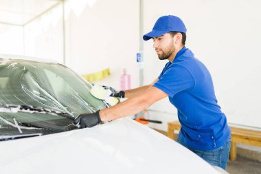 Latin bir adam araba yıkamada arabanın ön camını temizlemeye çalışıyor. Sabun ve araba camlarını silmek için sünger kullanan iyi görünümlü bir işçi.