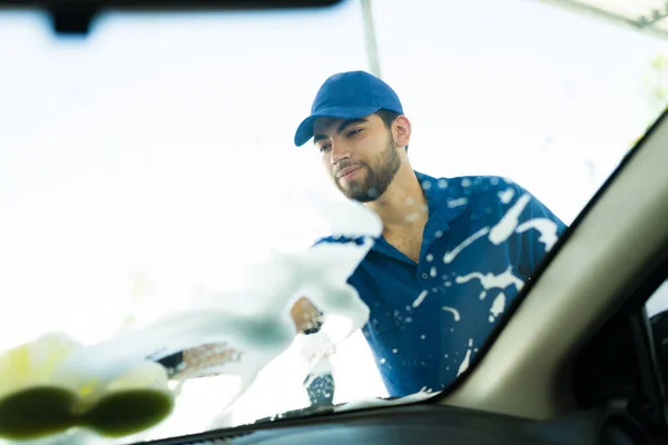 Happy to be doing a good job at the car wash. Handsome latin worker cleaning the car windshield with soap and a sponge