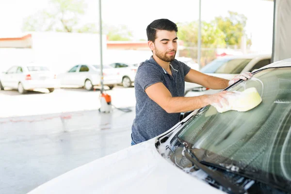 Handsome hispanic young man using soap and a sponge to wash the dirty windshield of his vehicle at a self service car wash