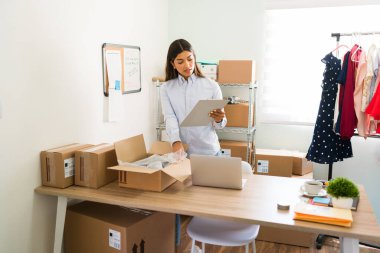 İş yerindeki çevrimiçi müşterilere paket göndermeye hazırlanırken kontrol listesine bakan çekici genç bir kadın.