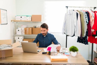 Odaklanmış işyeri sahibi ofis masasında oturuyor ve çevrimiçi e-ticaretinde ürünleri ve yeni moda kıyafetlerini güncelliyor. 