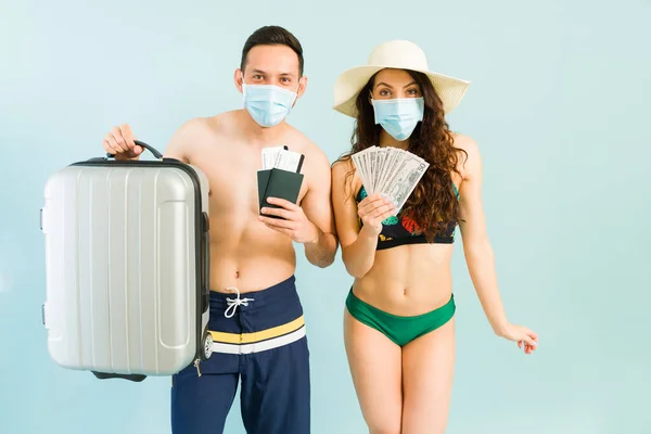 Getting ready to travel. Happy couple in swimwear and face masks holding their suitcase, money and plane tickets to a tropical beach