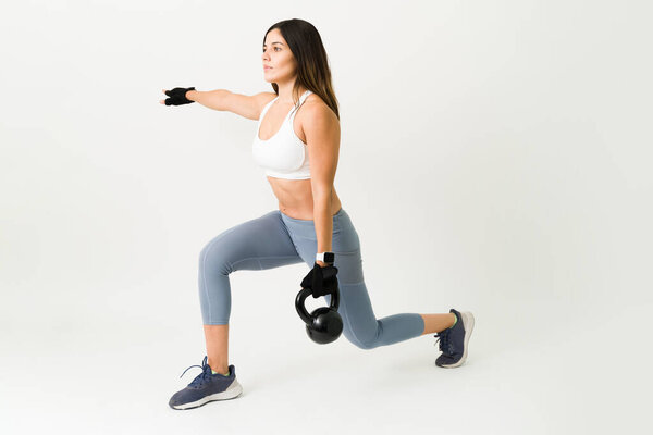 Beautiful sporty woman doing lunges while holding a kettlebell. Fit strong athlete exercising with weights for her daily training program