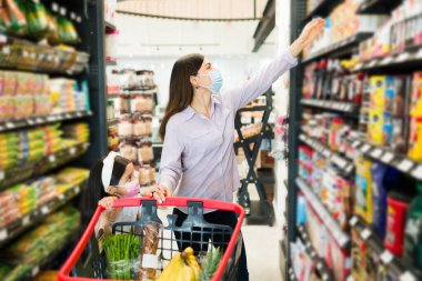 Çekici bir anne ve maske takan sevimli bir kız süpermarket koridorunda ürünleri seçiyor ve alışveriş arabasına bir şeyler koyuyor. 