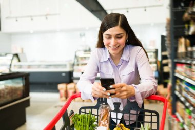 Süpermarkette mesajlaşıyoruz. Mutlu genç bir kadın mesaj atarken ya da markette sosyal medyayı kontrol ederken gülümsüyor. 