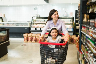 Neşeli anne ve sevimli kızı süpermarket koridorunda alışveriş arabasıyla oynarken gülümsüyorlar. 