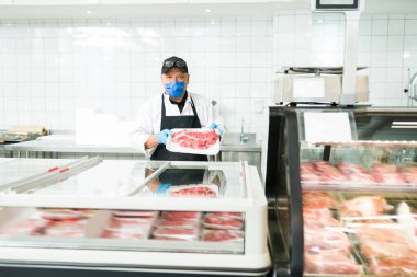 Kasapta çalışırken maske takan bir erkek satıcı. Süpermarkette çiğ et satan bir işçinin portresi. 