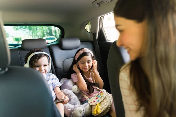 快乐的小男孩和小女孩坐在车里 带着耳机在平板电脑上看电影 一个带着可爱孩子开车的年轻女人 — 图库照片