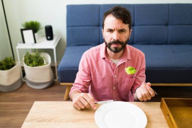 Portrait of an upset and depressed man with loss of appetite looking at his dinner plate and trying to eat lunch despite his eating disorder clipart