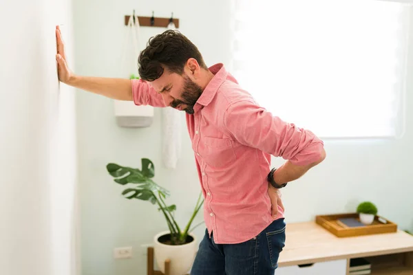 腰をこすり腰痛に苦しみながら リビングの壁に寄りかかって息をのむストレスの多い男 — ストック写真