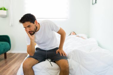 Pijamalı sinirli yakışıklı adam yatakta oturup uyuyan kız arkadaşıyla yaptığı kavgayı düşünüyor. 